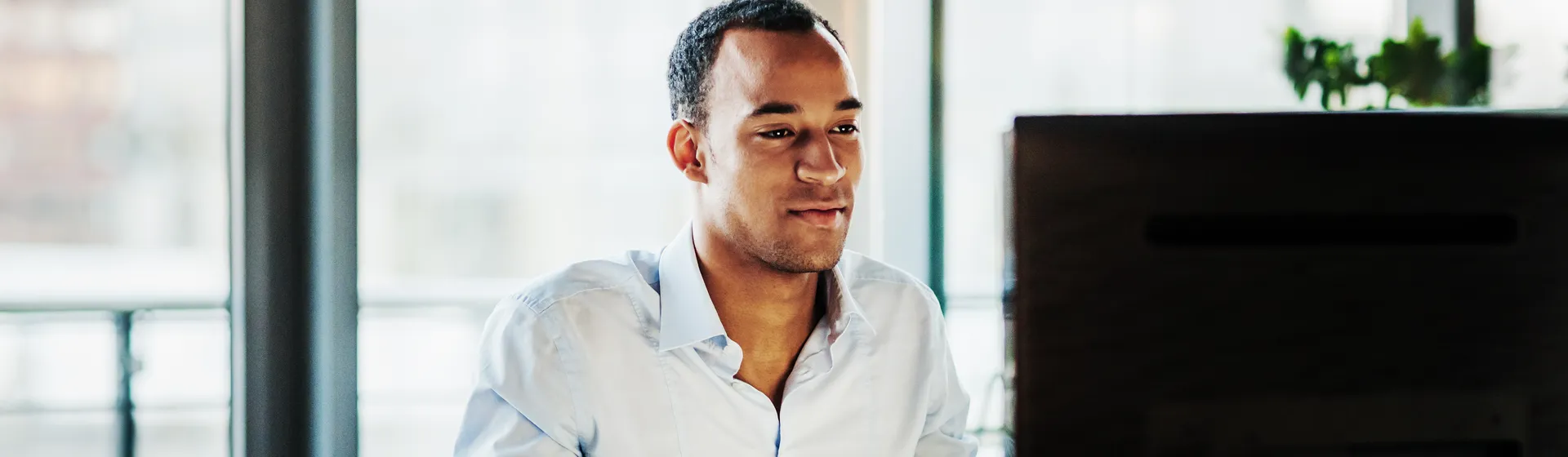 Un homme vêtu d'une chemise bleu clair est assis à un bureau et travaille avec concentration sur un ordinateur dans un bureau moderne.