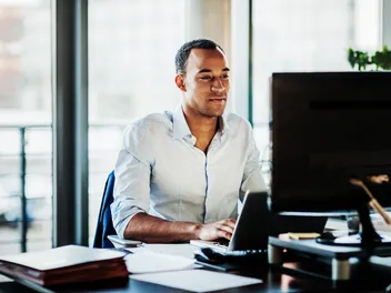 Un homme vêtu d’une chemise bleu clair travaille, concentré, sur un ordinateur dans un bureau moderne.