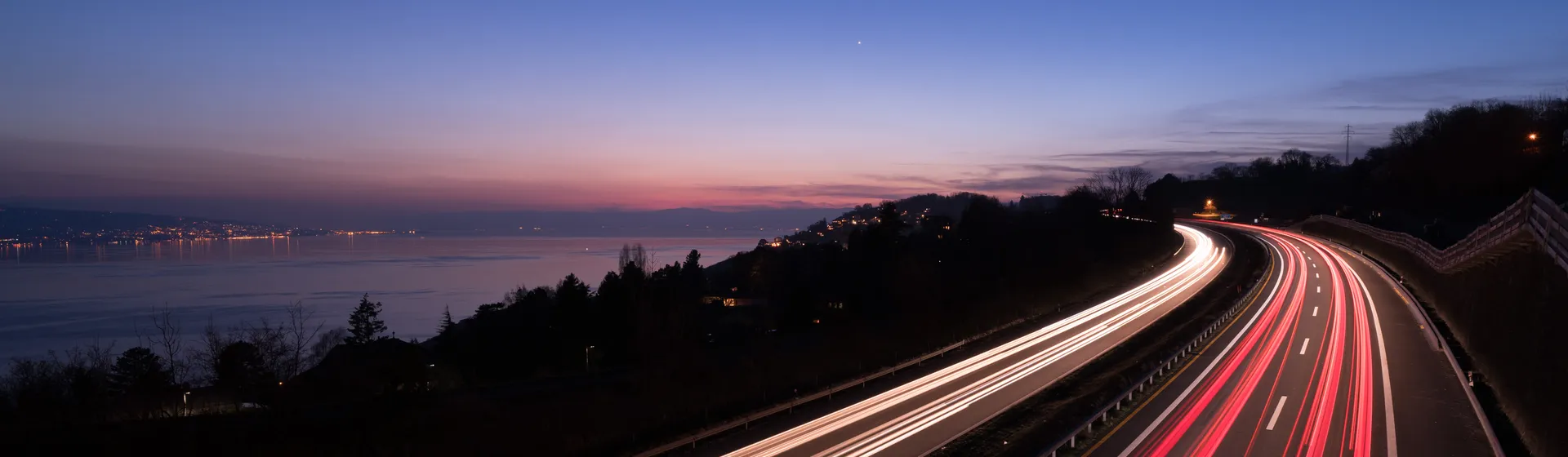 Eine kurvige Autobahn bei Dämmerung mit verschwommenen Lichtern vorbeifahrender Autos und einem ruhigen See im Hintergrund.