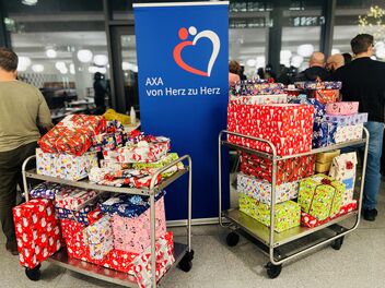 Zwei Wagen mit bunt verpackten Geschenken vor einem Banner von "AXA von Herz zu Herz" in einem belebten Raum.