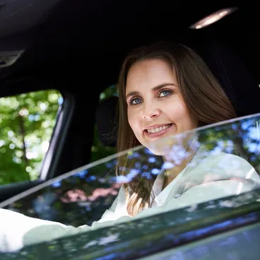 Eine Frau mit langen braunen Haaren lächelt, während sie in einem Auto sitzt und durch das offene Fenster auf der Fahrerseite schaut.