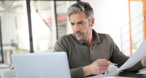 Un homme assis à son bureau regarde sérieusement son ordinateur portable, tenant des documents et un stylo dans ses mains.