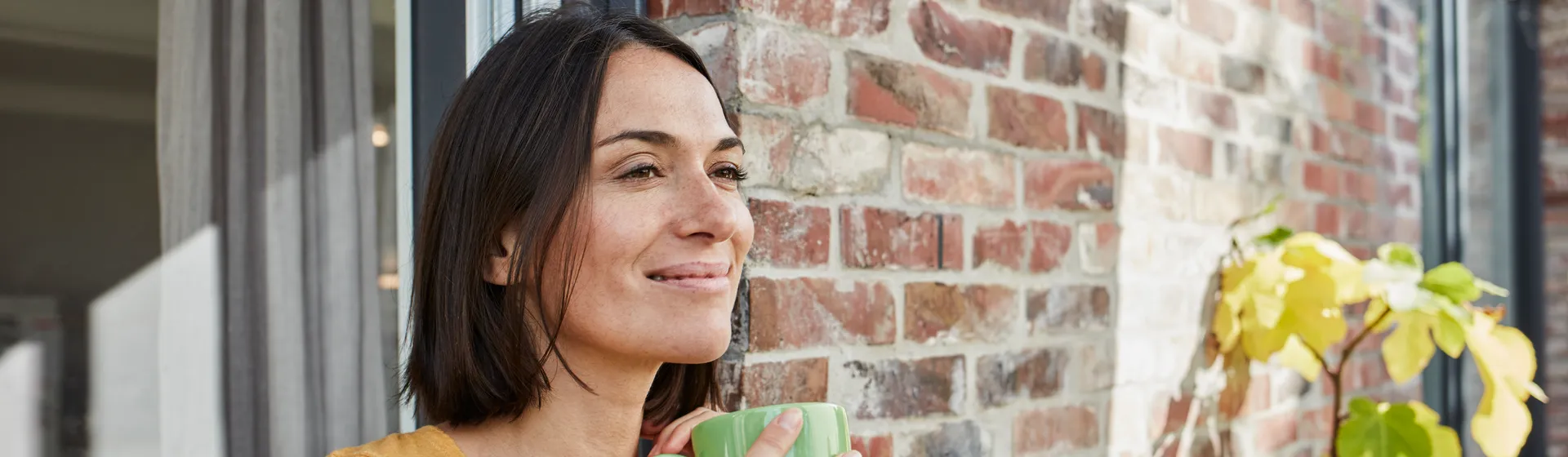 Junge Frau lehnt sich draussen am Fenster ihres Backsteinhauses an. Sie hält eine grüne Tasse in der Hand und lächelt.