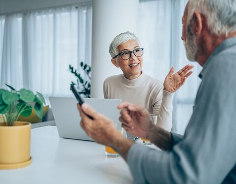 Une femme est assise à table chez elle. Elle a son ordinateur portable devant elle. Elle regarde son mari et lui explique quelque chose.