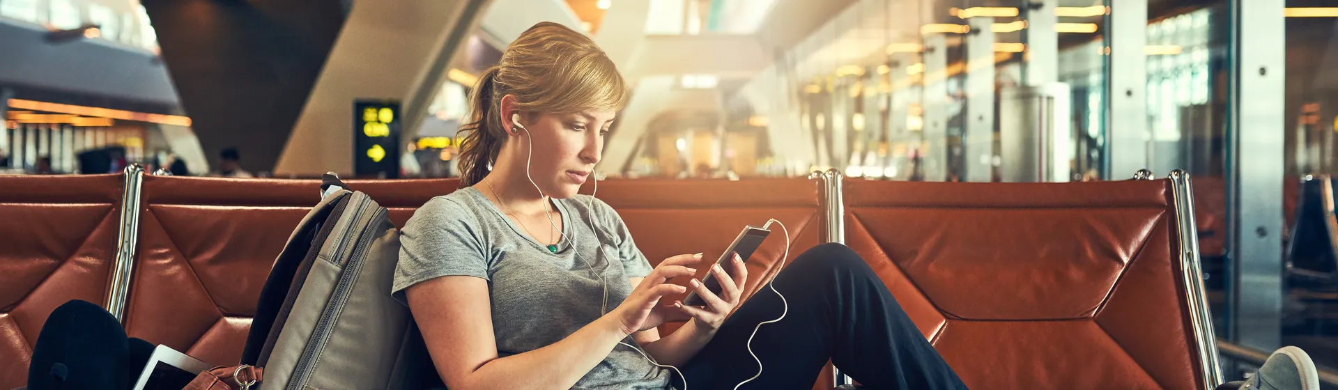 A woman is sitting relaxed on an airport seat, using her smartphone and has a rucksack and a coffee cup next to her.