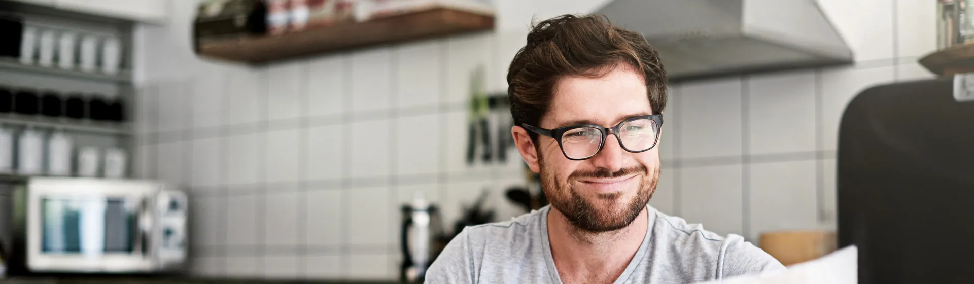 Giovane uomo con gli occhiali è seduto in cucina davanti al suo laptop e legge il giornale.