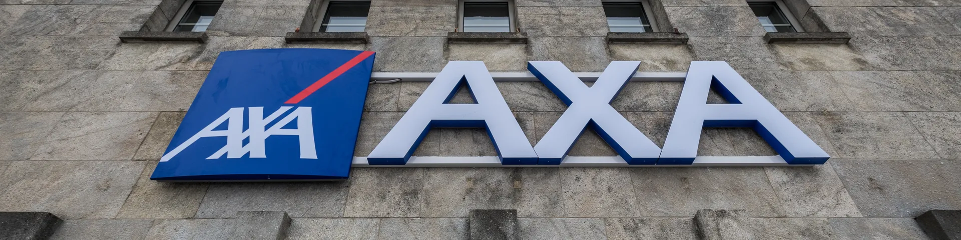 La photo montre le logo d’AXA Assurances sur un bâtiment gris avec plusieurs fenêtres à l’étage supérieur.