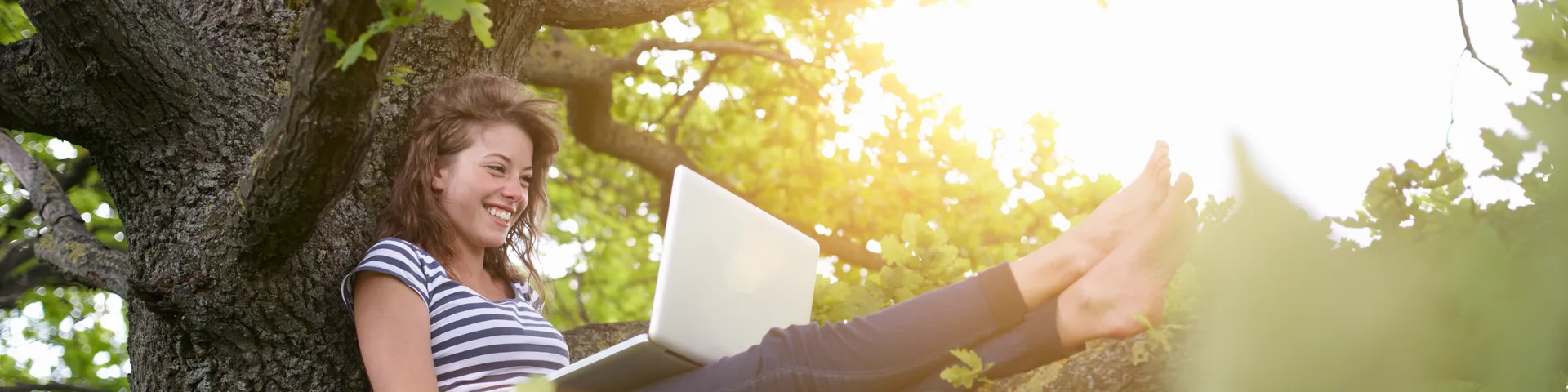 Une jeune femme souriante est assise dans un arbre et travaille sur un ordinateur portable à la lumière du soleil, entourée de feuilles vertes.