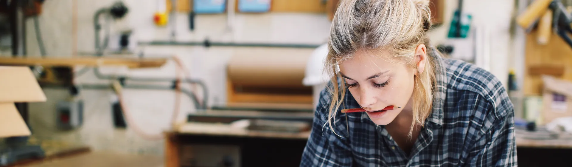 A woman in a checked shirt works intently on a drawing in a workshop, surrounded by tools and wooden materials.