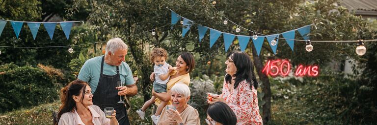 Une famille est assise et debout autour d'une table recouverte d'une nappe blanche dans le jardin. Ils rient et font la fête. Au-dessus d'eux, il y a une guirlande lumineuse et des triangles de fête bleus.