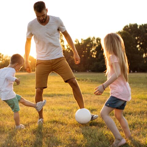 A father plays football with his son and daughter in a sunny field.