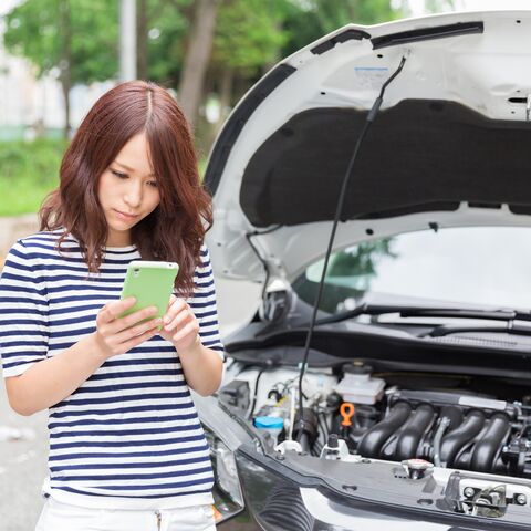 Una donna con una maglietta a righe è in piedi accanto a un'auto con il cofano aperto e guarda il suo cellulare come se stesse cercando aiuto.