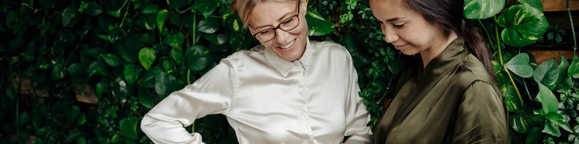 Zwei Frauen stehen vor einer grünen Blattwand, eine ältere Frau mit Brille und eine jüngere Frau in einem Gespräch vertieft und lächelnd.