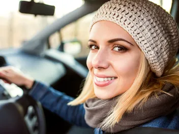 A young woman with blond hair and a knitted hat smiles out of the car window.