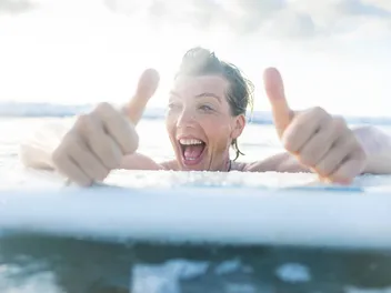 Une femme s’accroche à une planche de surf. Elle rit et lève les pouces.