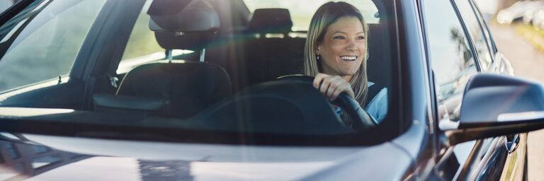 Eine lächelnde Frau mit blonden Haaren sitzt am Steuer eines Autos und blickt freudig nach vorne auf die Strasse. Die Aufnahme erfolgt durch die Windschutzscheibe und zeigt sie in einer hellen, sonnigen Umgebung.