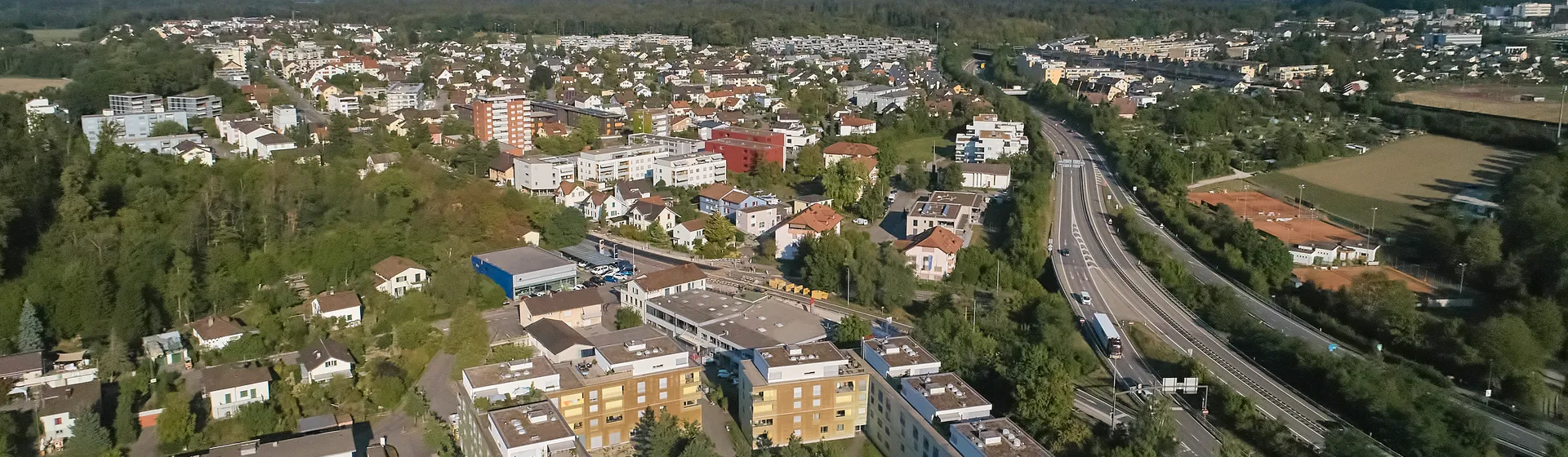 Foto aerea di una zona residenziale con diversi edifici residenziali, circondati da alberi e strade, e un’autostrada sullo sfondo.
