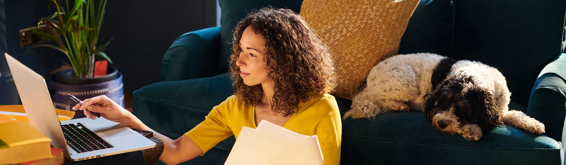 Une femme vêtue d'un T-shirt jaune est assise par terre devant un canapé turquoise. Elle tient un document dans la main gauche et un stylo dans la droite. Elle regarde un MacBook sur la table basse à sa gauche et un chien noir et blanc est couché sur le canapé à sa droite.