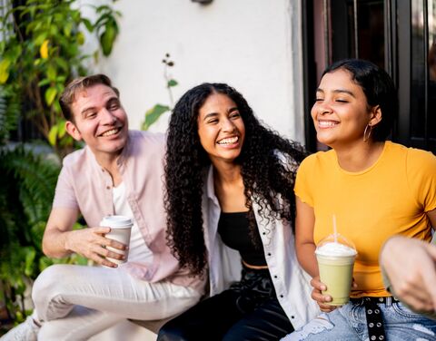 Drei lachende Freunde sitzen draussen auf einer Bank und geniessen Getränke; einer hält Kaffee, die anderen Smoothies, umgeben von Pflanzen.