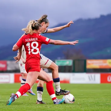 Deux joueuses de football se disputent le ballon pendant un match, avec en toile de fond des montagnes et des panneaux publicitaires.