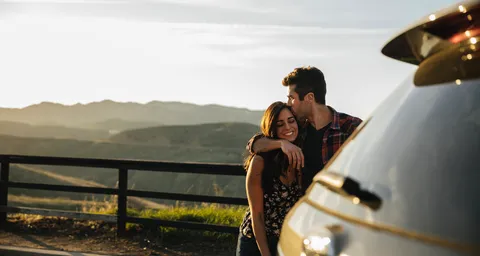 Ein Paar umarmt sich liebevoll neben einem Auto, mit einer malerischen Berglandschaft und einem Sonnenuntergang im Hintergrund.