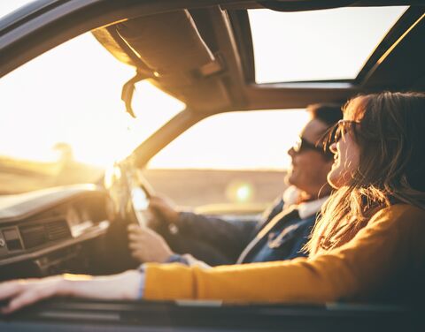 Eine lächelnde Frau mit Sonnenbrille sitzt im Auto, ihr Haar weht im Wind, während die Abendsonne durch das Fenster scheint.