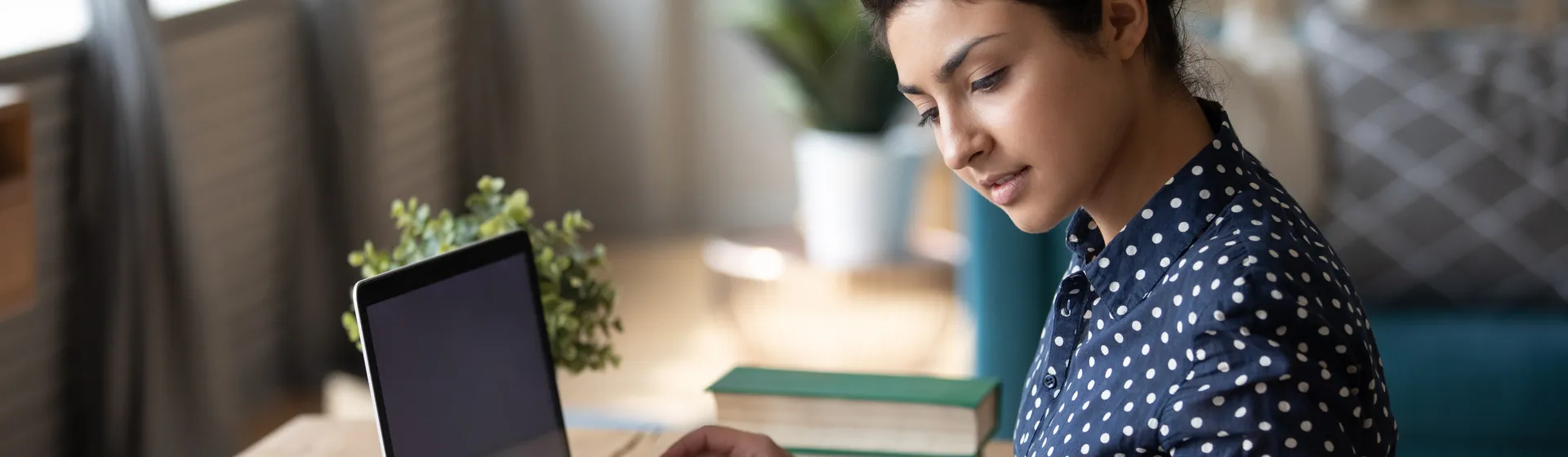 Une femme est assise à un bureau avec un ordinateur portable et étudie des documents, entourée de livres et de plantes dans une pièce lumineuse.