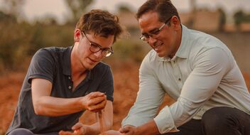 Two men with glasses kneel on red earth. One of them lets the red, dusty earth run through his fingers.