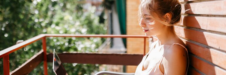 Une jeune femme détendue assise sur son balcon qui regarde son ordinateur portable.