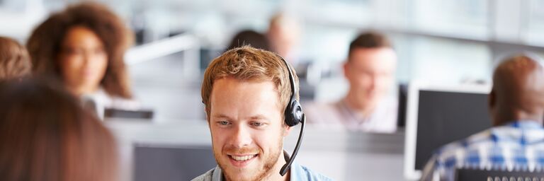 Ein junger Mann mit Headset lächelt und sitzt in einem modernen Büro, während er mit einer Person vor ihm kommuniziert.