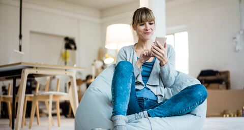 A woman sits on a bean bag, smiling as she taps out a message on her phone.