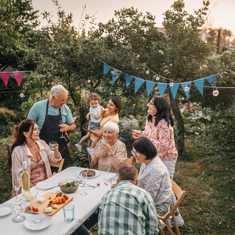 Famiglia seduta e in piedi intorno a un tavolo con una tovaglia bianca in giardino. Ridono e festeggiano. Sopra di loro sono appesi un filo di luci e triangoli blu da festa.