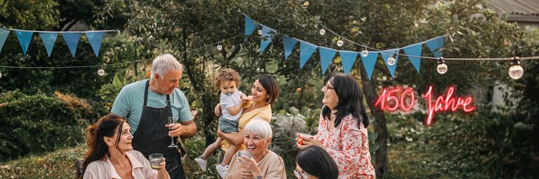 A family is gathered around a table with a white tablecloth in a garden. They are laughing and celebrating. String lights and blue bunting hang above them.