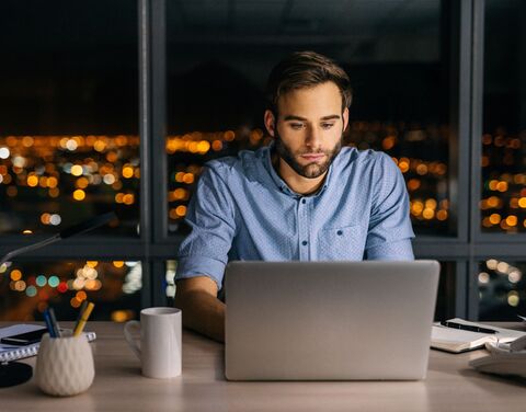 Un uomo dai capelli scuri in camicia blu è seduto di fronte al laptop della sua moderna postazione di lavoro. Grandi finestre sullo sfondo, in cui si intravedono riflesse le luci della città nel buio della sera.