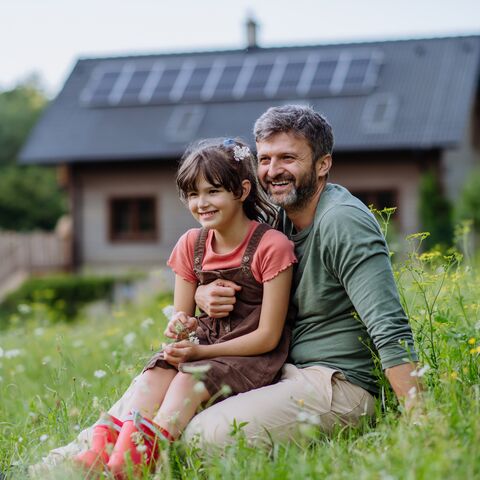 Ein Vater sitzt mit seiner Tochter auf einer Blumenwiese und beide schauen lächelnd in die Ferne. Im Hintergrund steht ihr Haus.