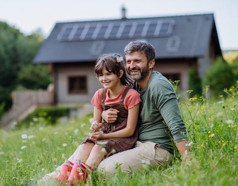 A father is sitting with his daughter in a flower meadow and both are looking into the distance with a smile. In the background is their house.