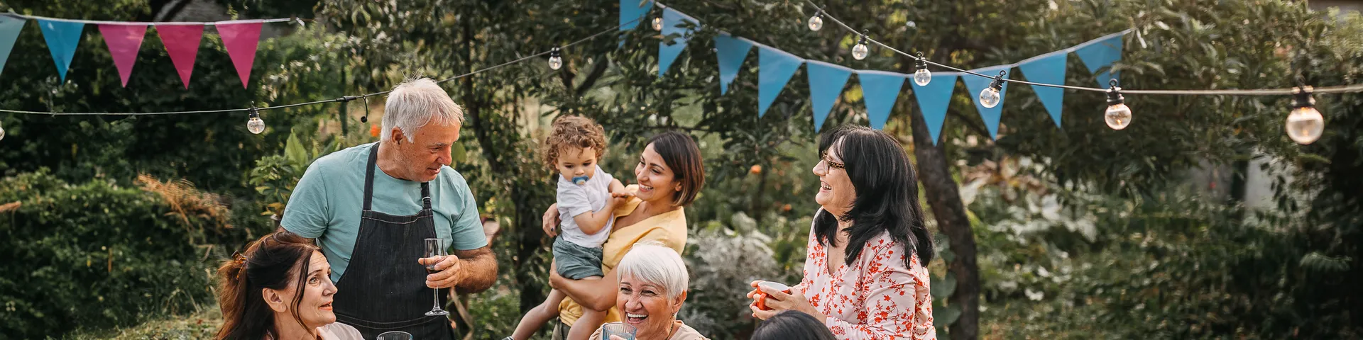 A family is gathered around a table with a white tablecloth in a garden. They are laughing and celebrating. String lights and blue bunting hang above them.
