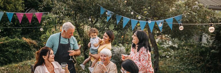 Family sitting and standing around a table with a white tablecloth in the garden. They are laughing and celebrating. A string of lights and blue party triangles hang above them.