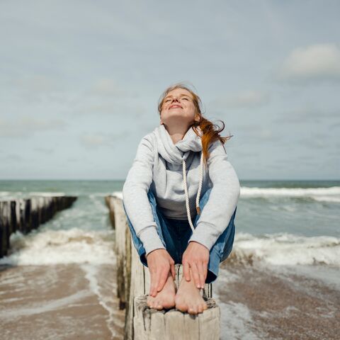 Frau mit weissem Pullover und roten Haaren sitzt auf einem Balken. Sie schaut mit geschlossenen Augen nach Oben und lächelt. Hinter ihr sieht man das Meer.