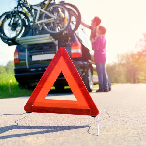 A red warning triangle stands on a country road in front of a car with bicycles mounted on its roof; two people stand in the background.