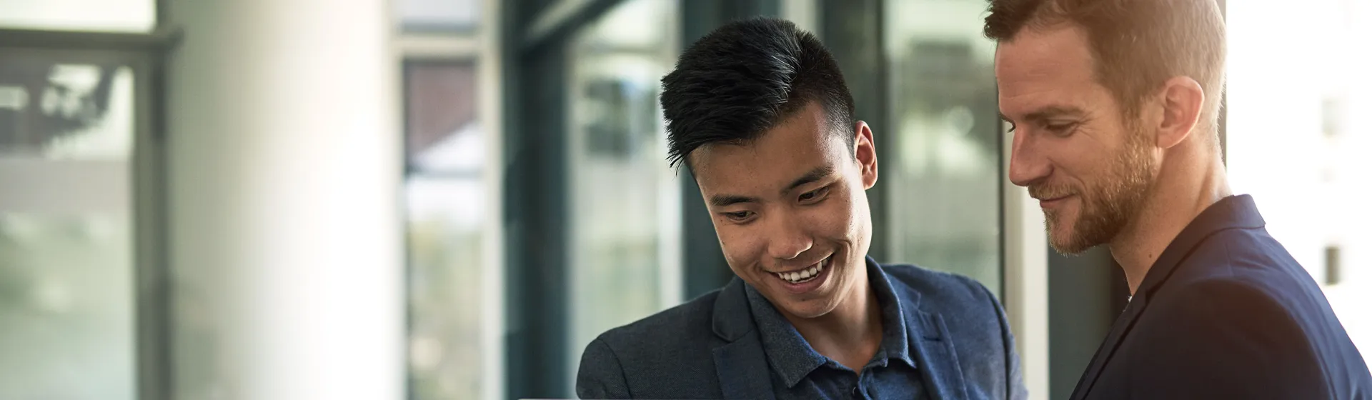 Deux hommes en tenue d'affaires se tiennent dans un bureau moderne et regardent en souriant un ordinateur portable que tient l'un d'eux.