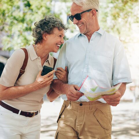 A smiling older couple outdoors, she holds a smartphone while he reads a map; both wear casual summer clothes.