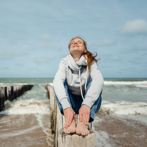 Eine rothaarige Frau mit hellem Kapuzenpulli und Jeans sitzt am Meer auf einem Holzpoller und genießt die Sonne.