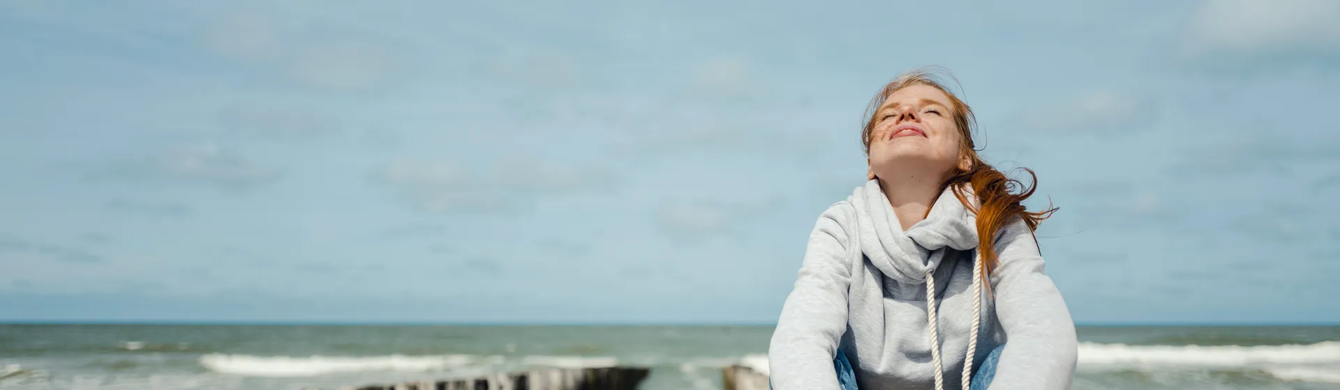 Une femme avec un pull blanc et des cheveux roux est assise sur une poutre. Elle regarde vers le haut, les yeux fermés, et sourit. Derrière elle, on voit la mer.
