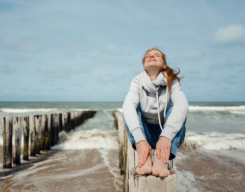 Woman with a white sweater and red hair sits on a beam. She looks upwards with her eyes closed and smiles. Behind her you can see the sea.