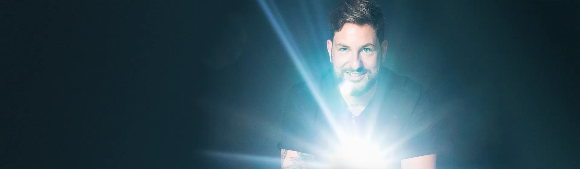 Un uomo con la barba sorride e guarda nella telecamera mentre una luce intensa brilla davanti a lui, lasciando lo sfondo scuro.