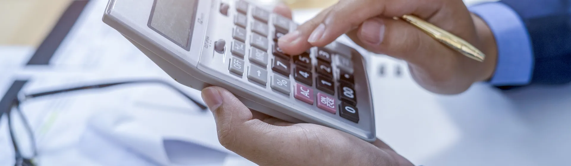 Une personne en tenue d'affaires utilise une calculatrice et tient un stylo doré ; des documents et des lunettes sont posés à l'arrière-plan.