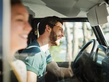 A smiling man drives a car while a laughing woman sits in the passenger seat. Sunlight shines through the window.