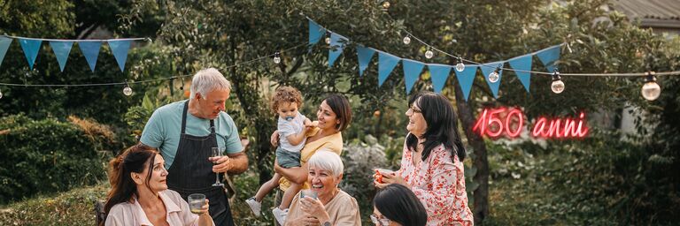 Famiglia seduta e in piedi intorno a un tavolo con una tovaglia bianca in giardino. Ridono e festeggiano. Sopra di loro sono appesi un filo di luci e triangoli blu da festa.