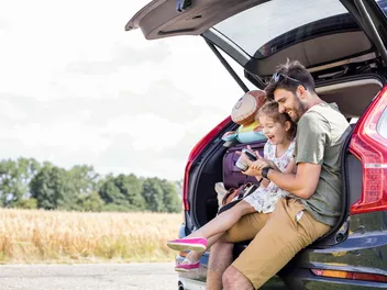 A father sits with his little daughter under the tailgate of a packed car. She looks at pictures on her father’s camera and laughs happily.